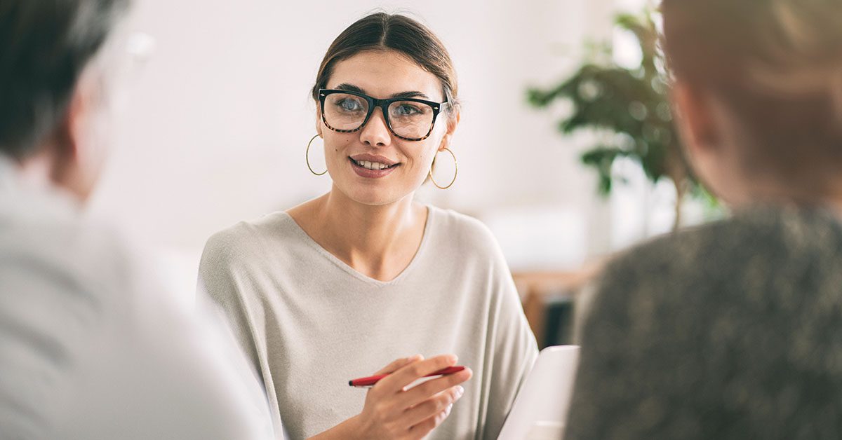 A professional financial advisor in glasses and a light sweater, smiling and discussing investment strategies with two clients in a modern office setting.