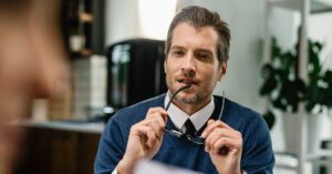 A thoughtful financial advisor plans the day ahead, holding glasses while reviewing priorities in a bright office.