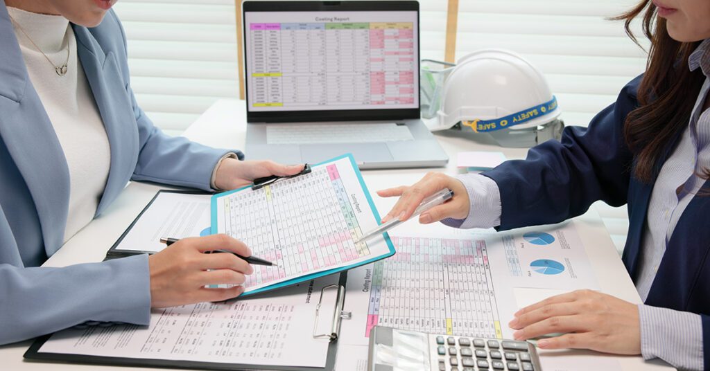 Two financial professionals review spreadsheets and planning documents together at a desk while analyzing workflow and operational data.